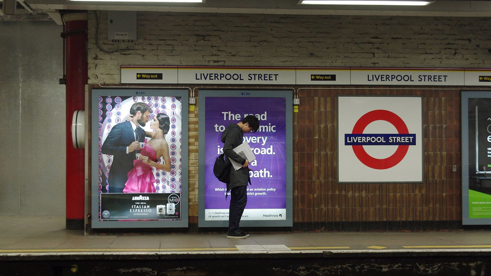 A lone commuter waiting on a London station platform beside two large backlit advertising panels